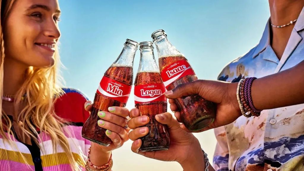 Tres personas de cerca, con ropa veraniega y pulseras, chocan tres botellas de Coca-Cola heladas y personalizadas en un día soleado. Las etiquetas dicen "Mia", "Logan" e "Isaac".